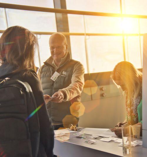 Lady at registration desk handing over a badge to a smiling man. on the right there is a young lady signing a paper.