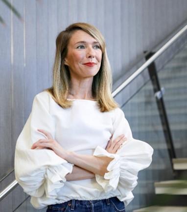 A professional headshot of Marika Nyyssönen, the ESG and Sustainability Manager at Hoas. She is standing, wearing a white blouse, smiling slightly, looking towards the right. Behind her is a staircase in a building