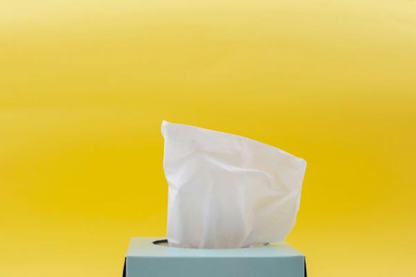 Picture of a light blue tissue box, with a tissue pulled up, on a bright yellow uniform background.